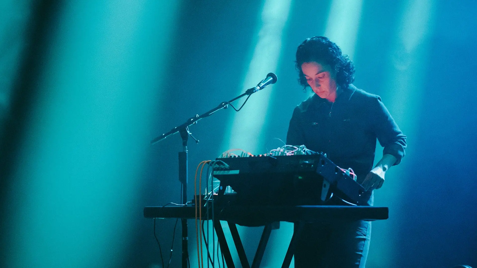 Su Shaw performing on stage with synth and microphone, warm blue lights