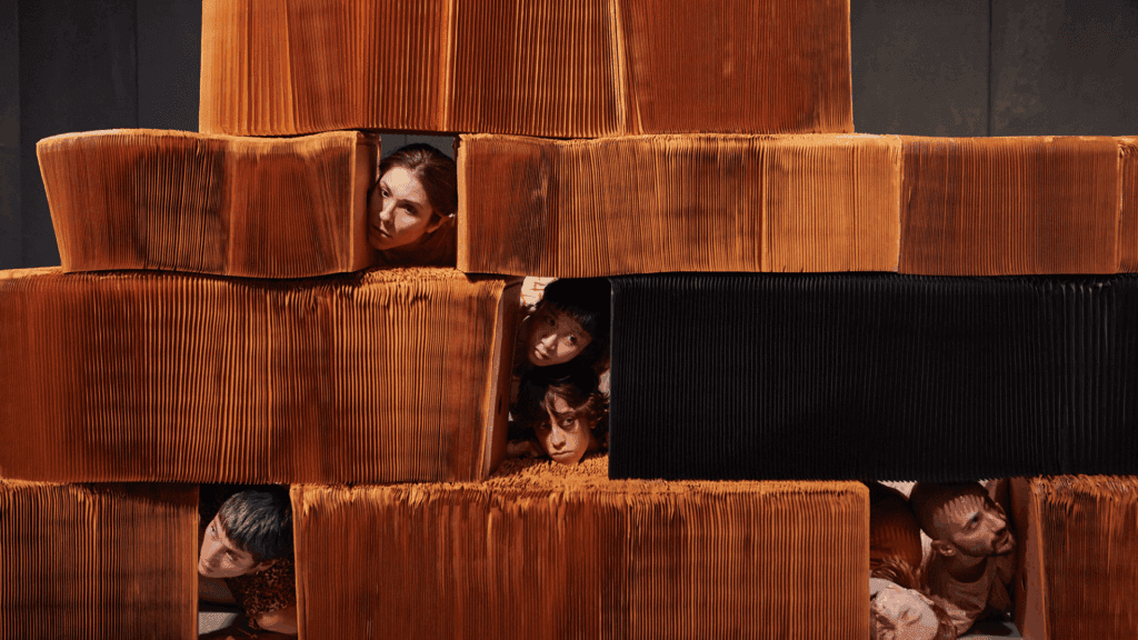 a group of people poke their faces through a stack of stylised orange papers that resemble a haybale
