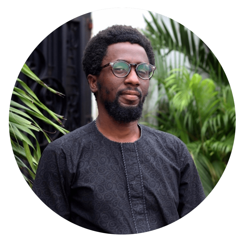 A black man with short hair, glasses and a beard looks at the camera. There are palm trees behind him.