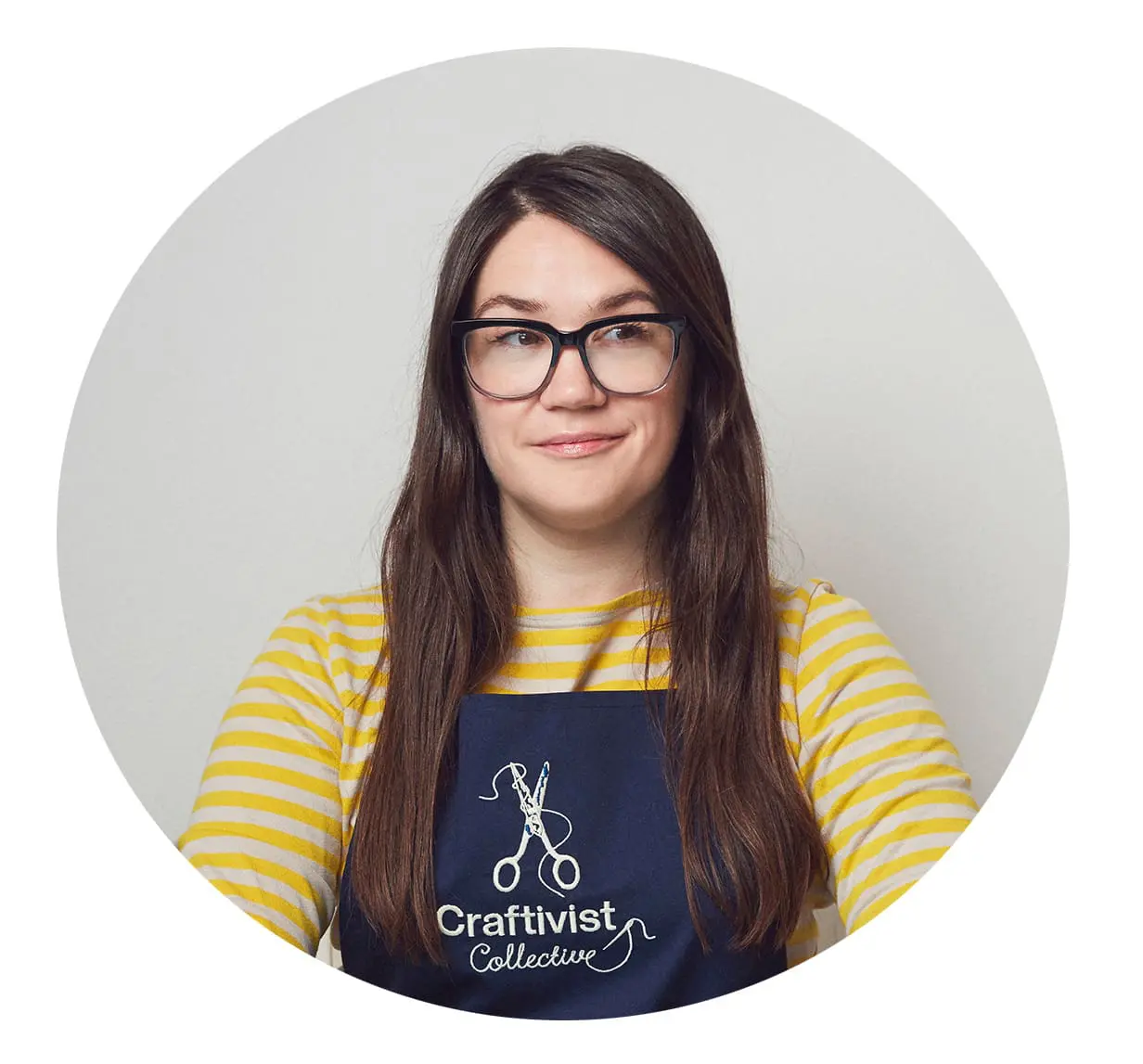A woman with long hair and glasses wearing an apron that says Craftivist Collective with a pair of scissors stitched above it. She is smiling.