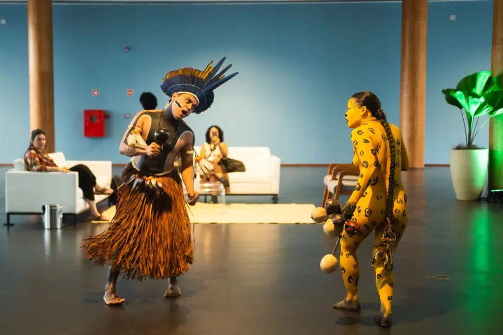 2 Indigenous artists dancing, with paint covered bodies and traditional attire in an indoor gallery space