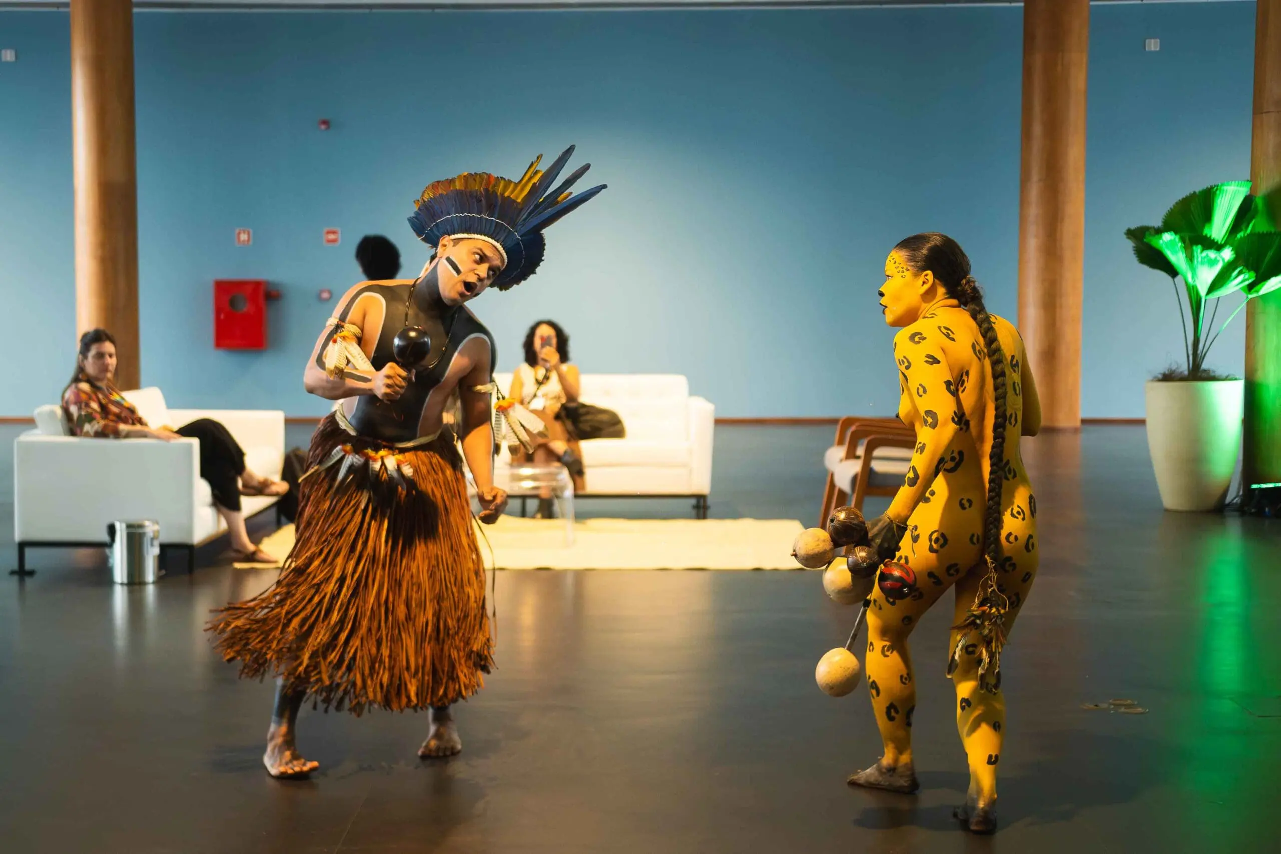2 Indigenous artists dancing, with paint covered bodies and traditional attire in an indoor gallery space