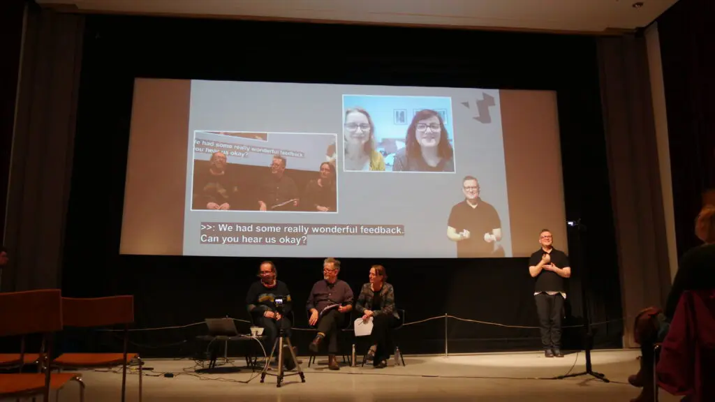 people on stage giving a presentation, there are some who have joined remotely and are online on the screen