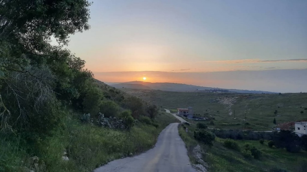 A path leads down to a small collection of buildings with the sun setting in the distance.