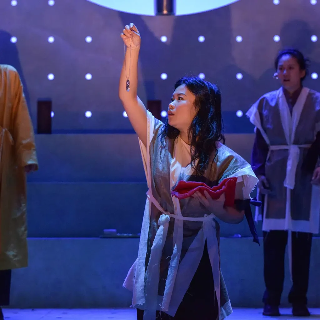 actor holding up a pendant to a light, looking shocked, and facing off to the side of the stage